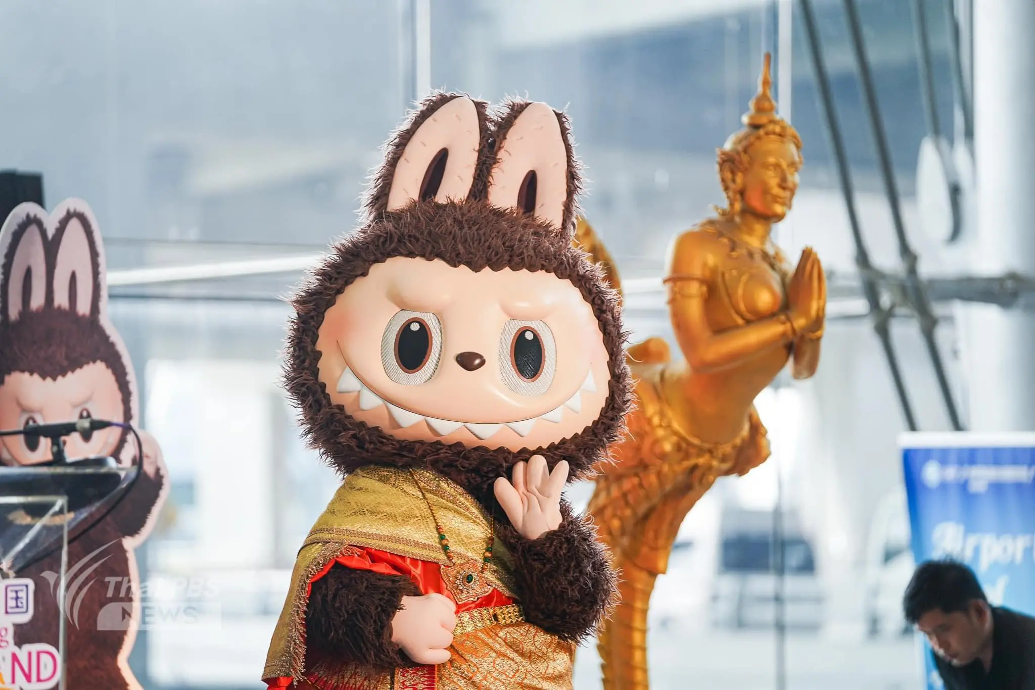 Life-size Labubu mascot wearing traditional Thai costume, posing at a public event with a golden statue in the background.