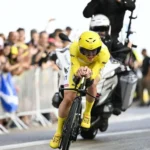 Cyclist in yellow jersey leading a time trial stage at Tour de France 2025, surrounded by cheering fans and team support vehicles.