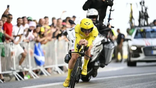 Cyclist in yellow jersey leading a time trial stage at Tour de France 2025, surrounded by cheering fans and team support vehicles.