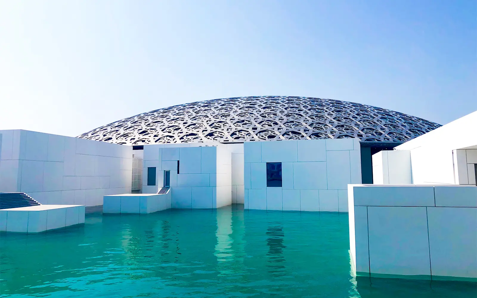 The stunning geometric dome of Louvre Abu Dhabi creates a serene reflection on the turquoise waters surrounding the museum.