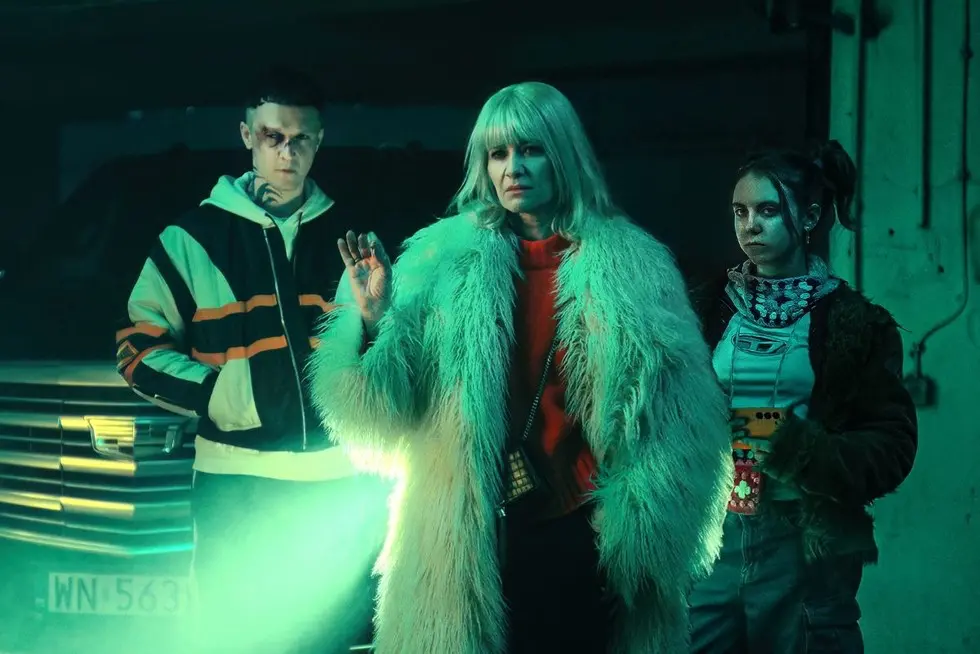 Aniela wearing a fur coat stands confidently between two edgy young characters in a dark garage, lit by eerie green lighting – scene from Netflix’s Polish series Aniela.