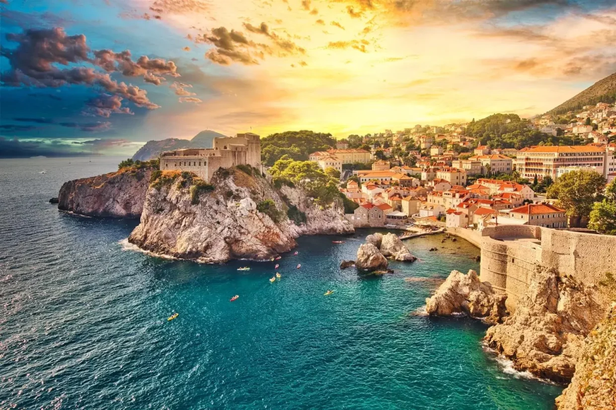 Scenic aerial view of Dubrovnik Old Town and fortress at sunset, with kayaks in the turquoise Adriatic Sea and dramatic clouds above the coastline.