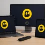 CyberGhost VPN interface displayed on multiple devices including smartphone, laptop, desktop monitor, tablet, TV remote, and router—all on a wooden desk.