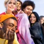 A diverse group of smiling Gen Z friends wearing colorful streetwear, standing close together outdoors on a sunny day, radiating youthful energy and positivity.