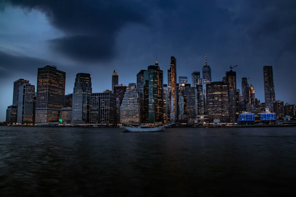 Stunning night view of Manhattan’s skyline with illuminated skyscrapers and a sailboat on the East River under dramatic clouds.