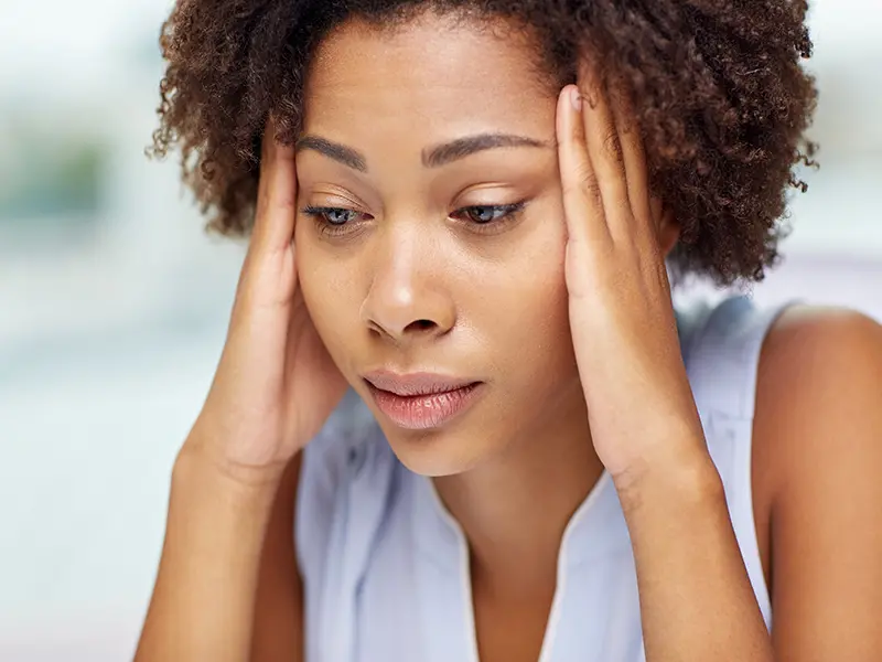 Woman holding her head with both hands, appearing fatigued and in pain—suggesting symptoms of a migraine or intense headache.