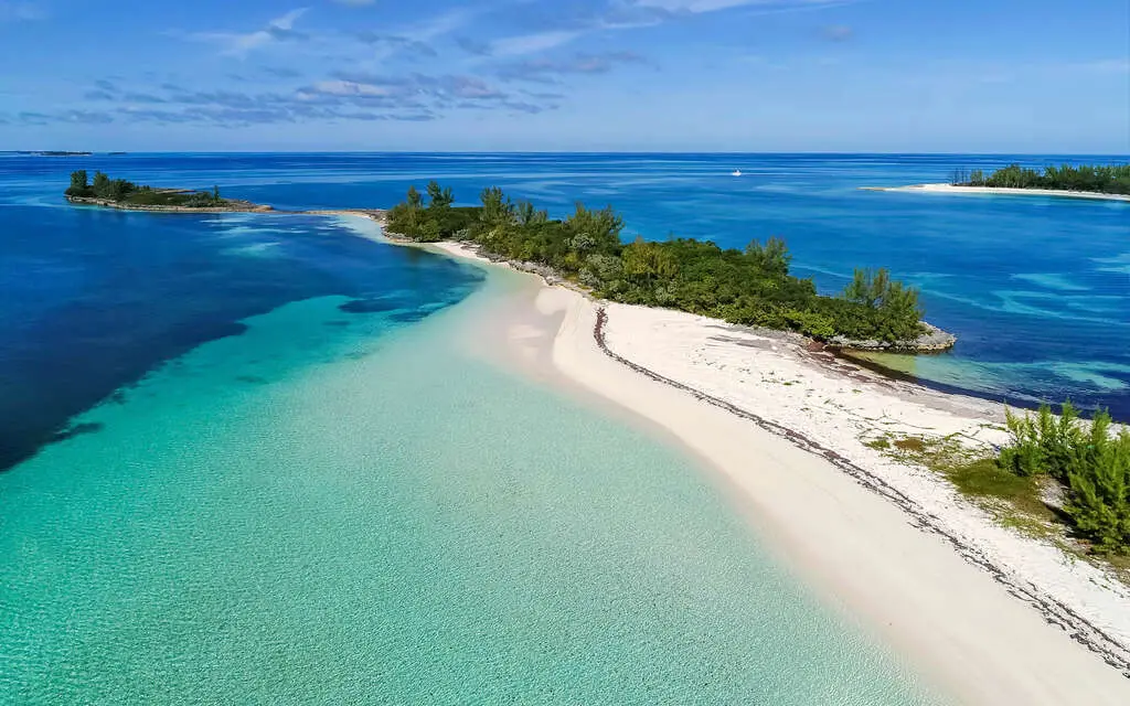 Aerial view of a pristine white sand beach and turquoise waters on a tropical island in the Bahamas, surrounded by lush greenery and calm ocean bays.
