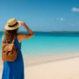 A woman in a blue dress and straw hat with a leather backpack stands on a tropical beach in the Bahamas, looking out at turquoise waters and distant islands.