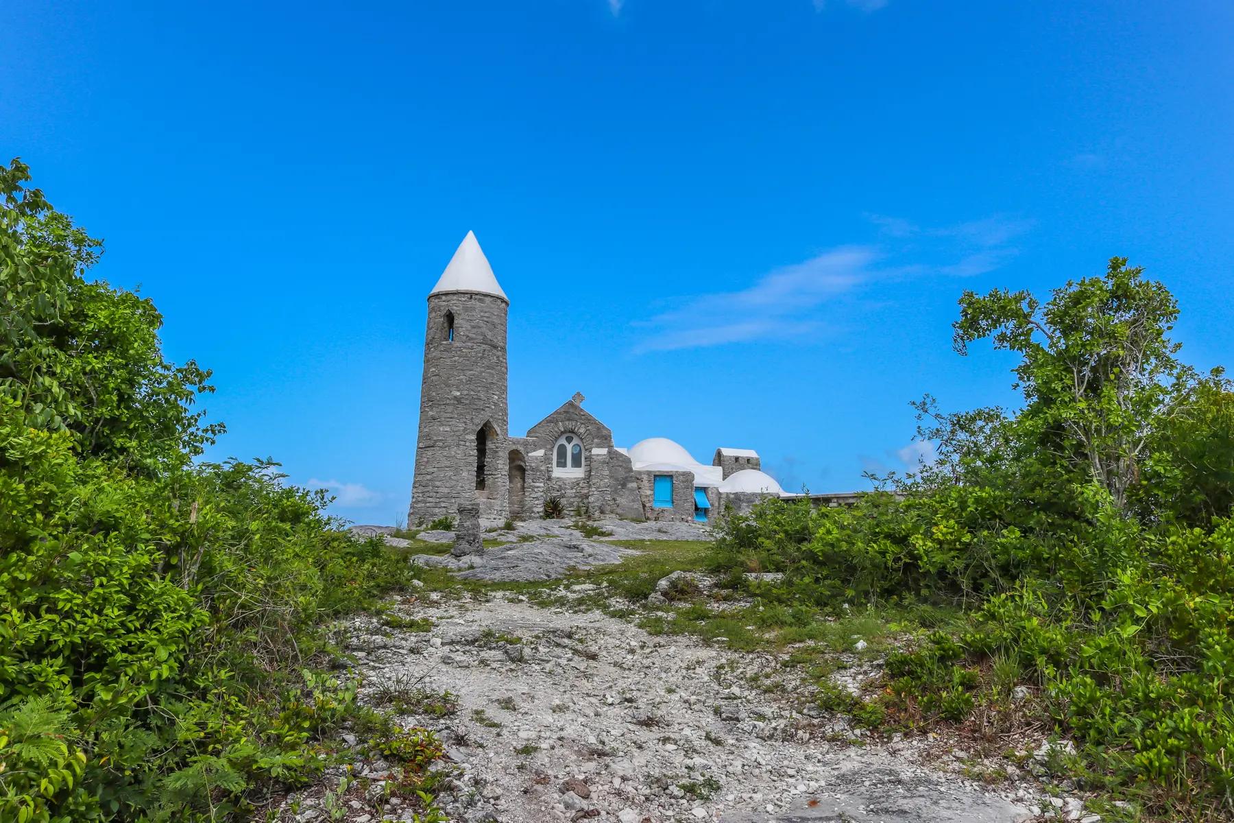 Scenic path leading to Mount Alvernia monastery on Cat Island, Bahamas, with stone tower, white domes, and lush tropical greenery under a clear blue sky.