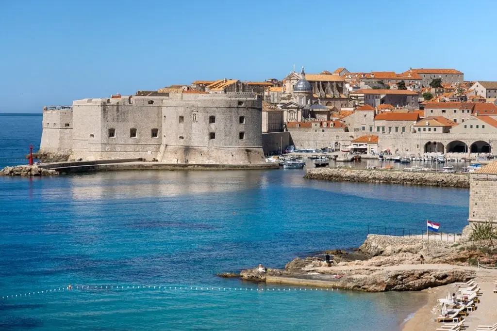 View of Dubrovnik’s Old Town walls and harbor with turquoise Adriatic waters, historic stone buildings, and red-tiled rooftops under a clear blue sky.
