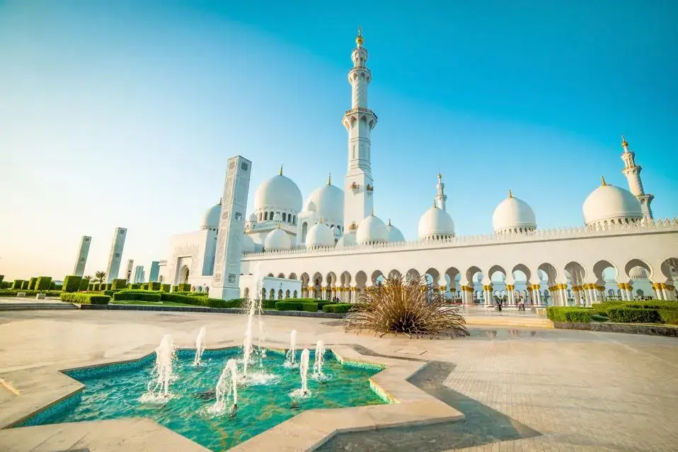 Exterior view of Sheikh Zayed Grand Mosque with fountains and white marble domes at sunset.