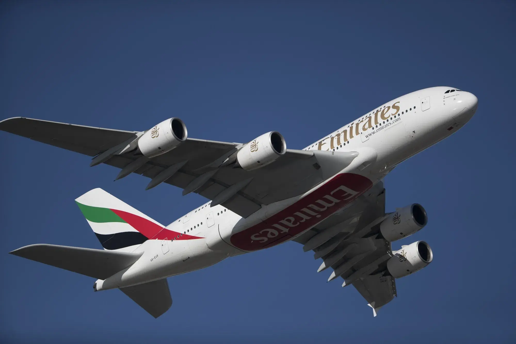 Close-up view of an Emirates Airbus A380 airplane taking off against a clear blue sky, showing the aircraft’s engines, logo, and tail fin with colorful stripes.