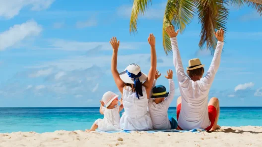 A happy family on vacation enjoys a perfect beach day, sitting together in the sand with their arms raised to celebrate the beautiful view.