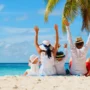 A happy family on vacation enjoys a perfect beach day, sitting together in the sand with their arms raised to celebrate the beautiful view.