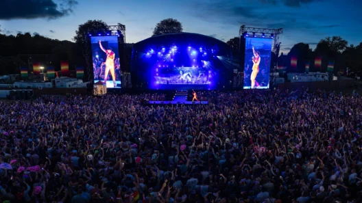 Massive crowd enjoying a vibrant nighttime performance at Brighton & Hove Pride, with a brightly lit stage, rainbow flags, and a performer in an orange outfit shown on giant screens.
