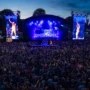 Massive crowd enjoying a vibrant nighttime performance at Brighton & Hove Pride, with a brightly lit stage, rainbow flags, and a performer in an orange outfit shown on giant screens.