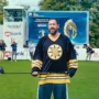 Adam Sandler as Happy Gilmore, standing confidently on a golf course in a Boston Bruins jersey, ready to swing, surrounded by cameras and spectators at a tournament.