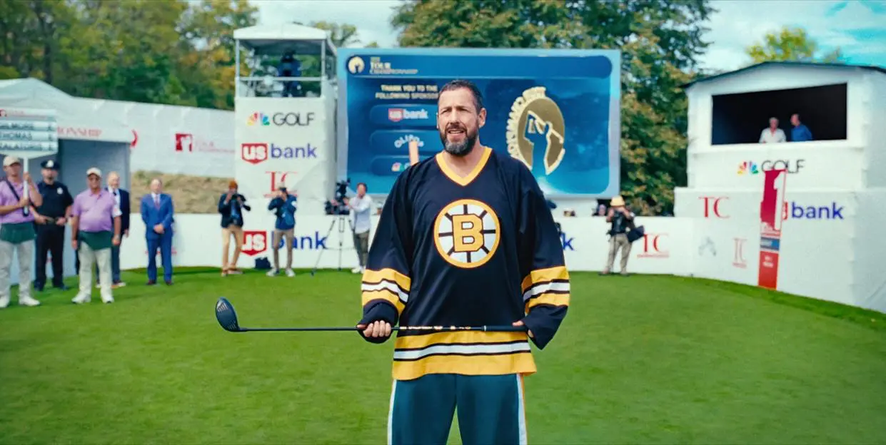 Adam Sandler as Happy Gilmore, standing confidently on a golf course in a Boston Bruins jersey, ready to swing, surrounded by cameras and spectators at a tournament.