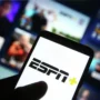 A hand in the foreground holds a smartphone with the ESPN+ logo lit up on its display, against a blurred background showing a screen with video content.