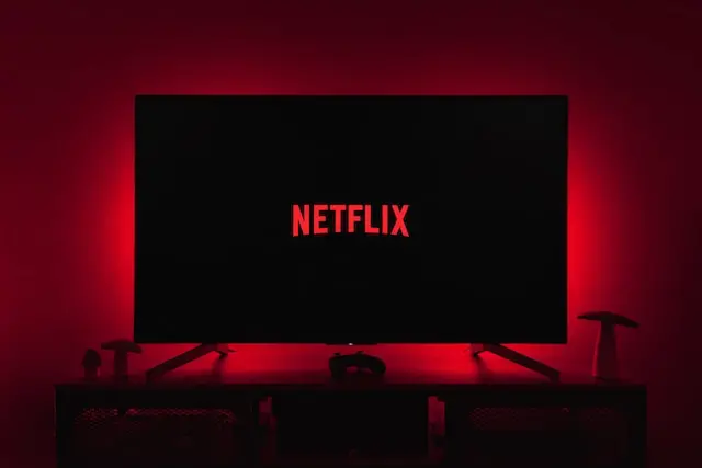 A dark room with a TV screen showing the Netflix logo, illuminated by a red backlight, representing night-time streaming and a cinematic home viewing experience.