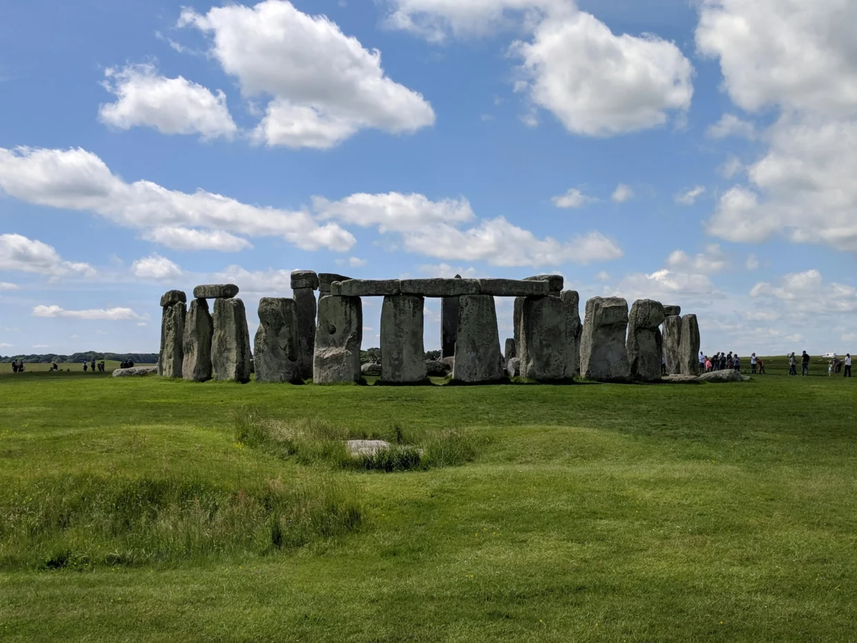 View of Stonehenge on a sunny day in Wiltshire, England, with clear skies, green grass, and tourists walking in the distance.