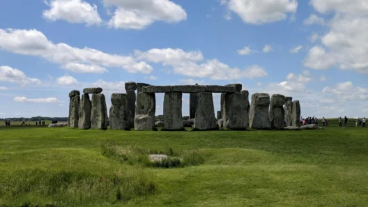 View of Stonehenge on a sunny day in Wiltshire, England, with clear skies, green grass, and tourists walking in the distance.