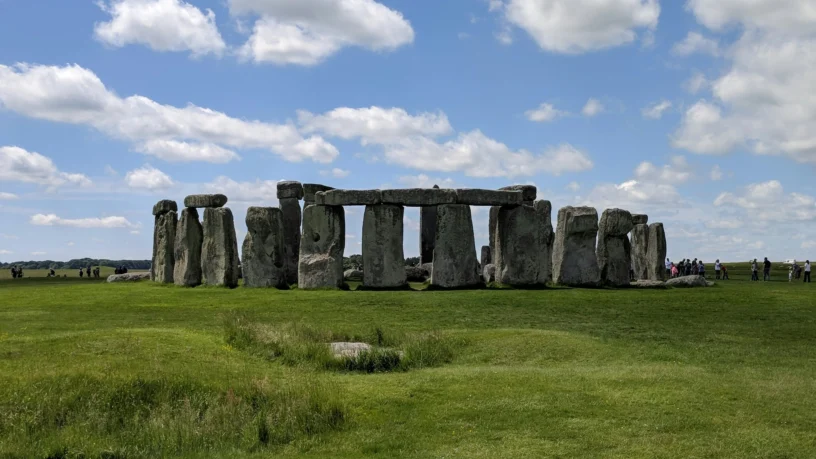 View of Stonehenge on a sunny day in Wiltshire, England, with clear skies, green grass, and tourists walking in the distance.