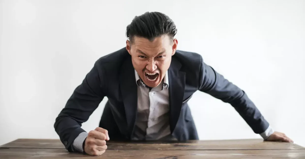 Angry man in a suit shouting and slamming his fist on a wooden table, expressing intense frustration or aggressive authority.