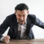 Angry man in a suit shouting and slamming his fist on a wooden table, expressing intense frustration or aggressive authority.