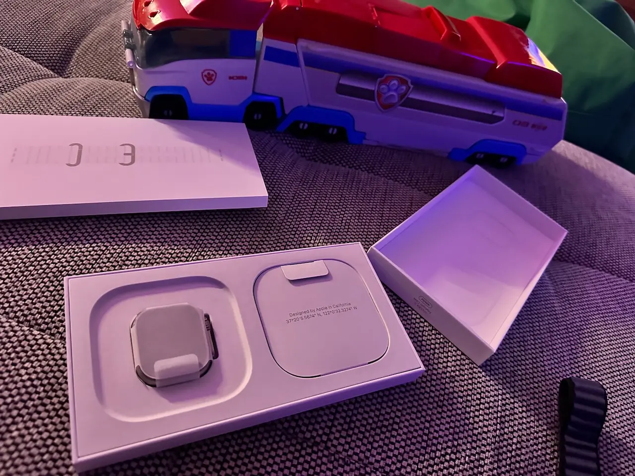 Apple Watch Ultra in its original box next to a Paw Patrol fire truck toy on a grey sofa.