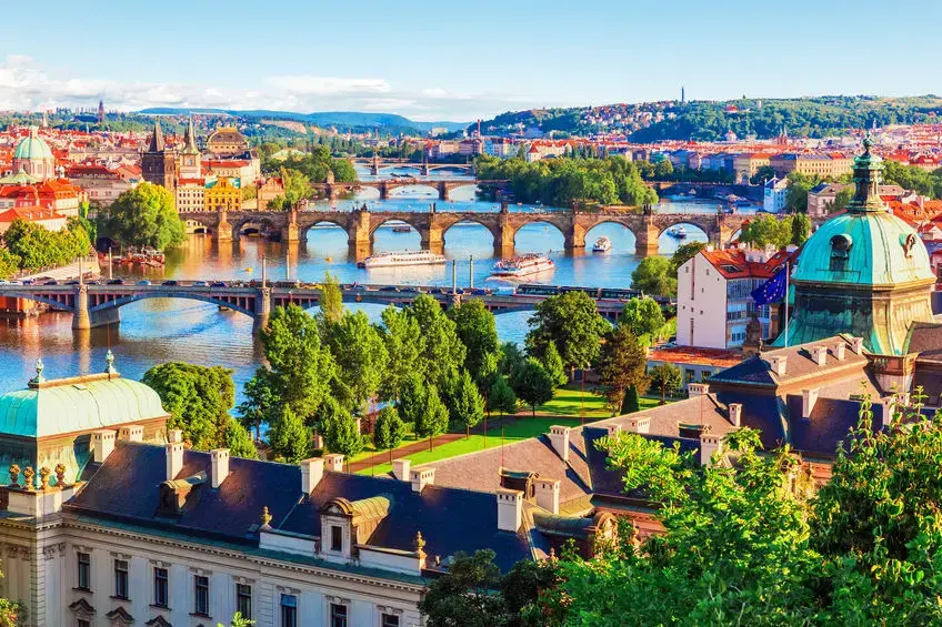 Scenic view of Prague with multiple historic bridges crossing the Vltava River, surrounded by colorful buildings, trees, and iconic architecture on a sunny day.