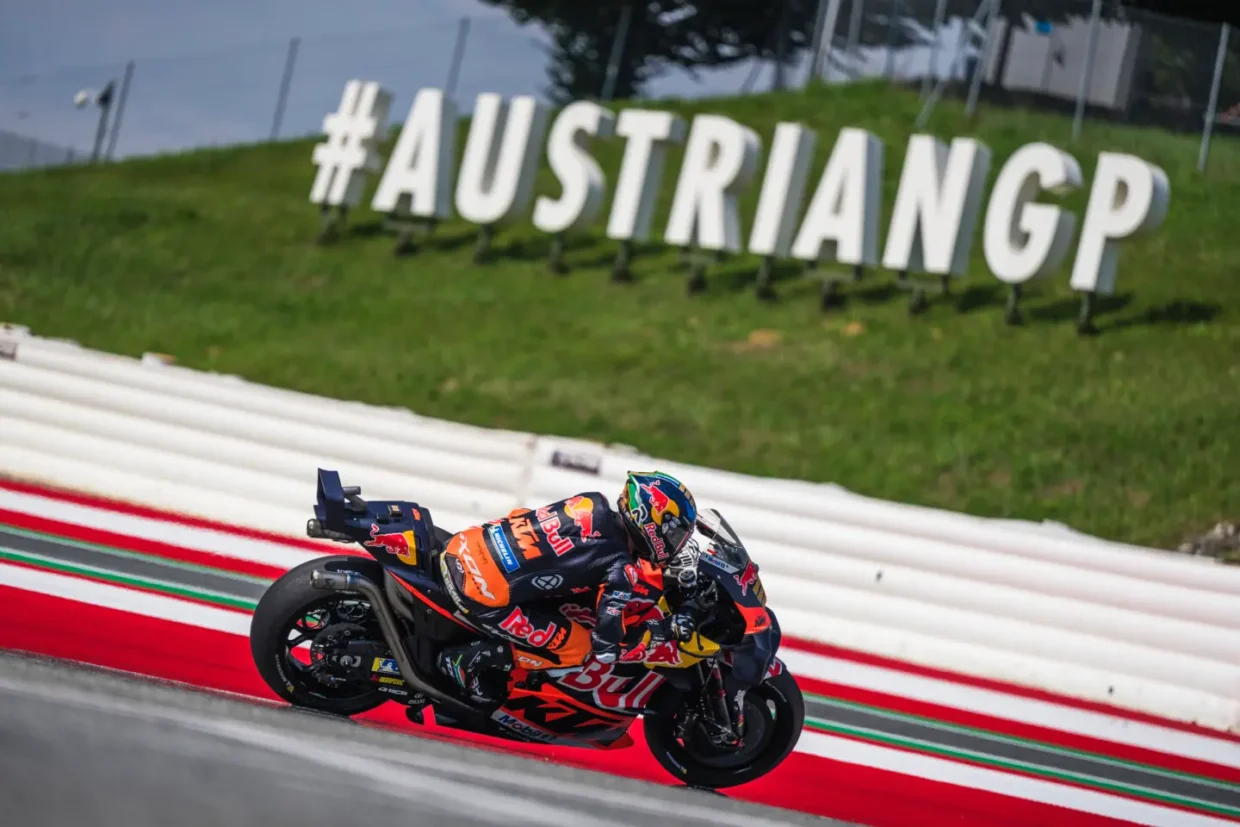 MotoGP rider on a KTM bike speeding through a corner at the Red Bull Ring, with the large #AUSTRIANGP sign in the background during the Austrian MotoGP 2025.