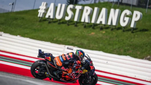 MotoGP rider on a KTM bike speeding through a corner at the Red Bull Ring, with the large #AUSTRIANGP sign in the background during the Austrian MotoGP 2025.