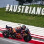 MotoGP rider on a KTM bike speeding through a corner at the Red Bull Ring, with the large #AUSTRIANGP sign in the background during the Austrian MotoGP 2025.