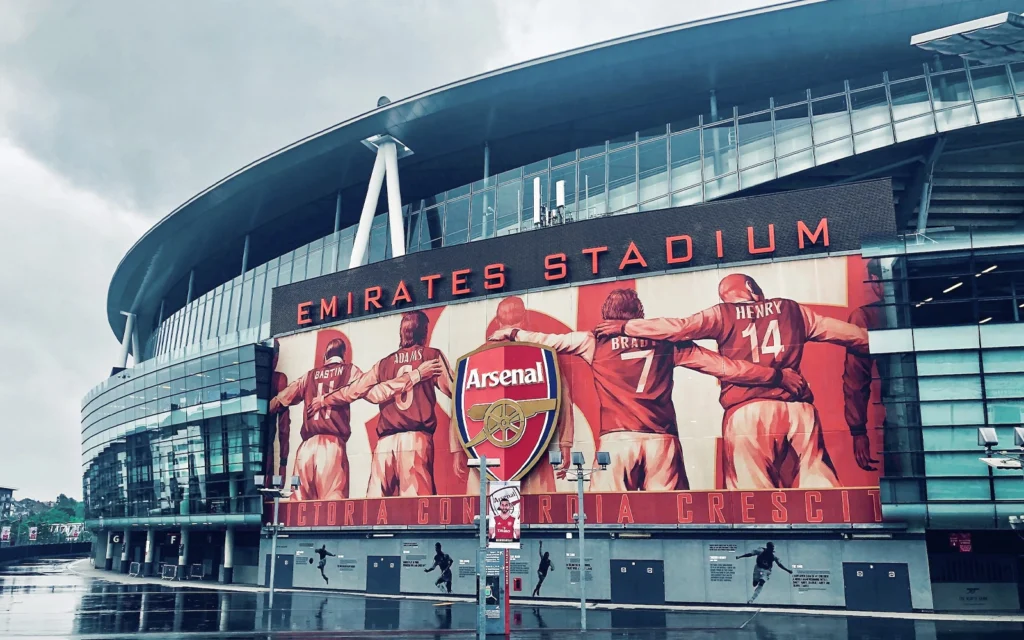 Emirates Stadium on a rainy day, featuring the iconic Arsenal crest and a mural of legendary players including Bastin, Adams, Brady, and Henry. This modern home of Arsenal FC showcases the club’s rich history and is the heart of every Gooner’s pilgrimage to North London.