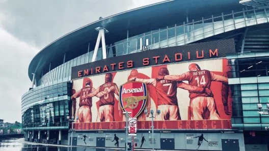Emirates Stadium on a rainy day, featuring the iconic Arsenal crest and a mural of legendary players including Bastin, Adams, Brady, and Henry. This modern home of Arsenal FC showcases the club’s rich history and is the heart of every Gooner’s pilgrimage to North London.