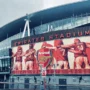 Emirates Stadium on a rainy day, featuring the iconic Arsenal crest and a mural of legendary players including Bastin, Adams, Brady, and Henry. This modern home of Arsenal FC showcases the club’s rich history and is the heart of every Gooner’s pilgrimage to North London.