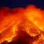 A dramatic eruption of an active volcano at night, with bright orange lava flows streaming down the slopes and thick clouds of glowing ash and smoke rising into the dark sky.