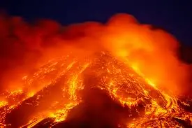 A dramatic eruption of an active volcano at night, with bright orange lava flows streaming down the slopes and thick clouds of glowing ash and smoke rising into the dark sky.