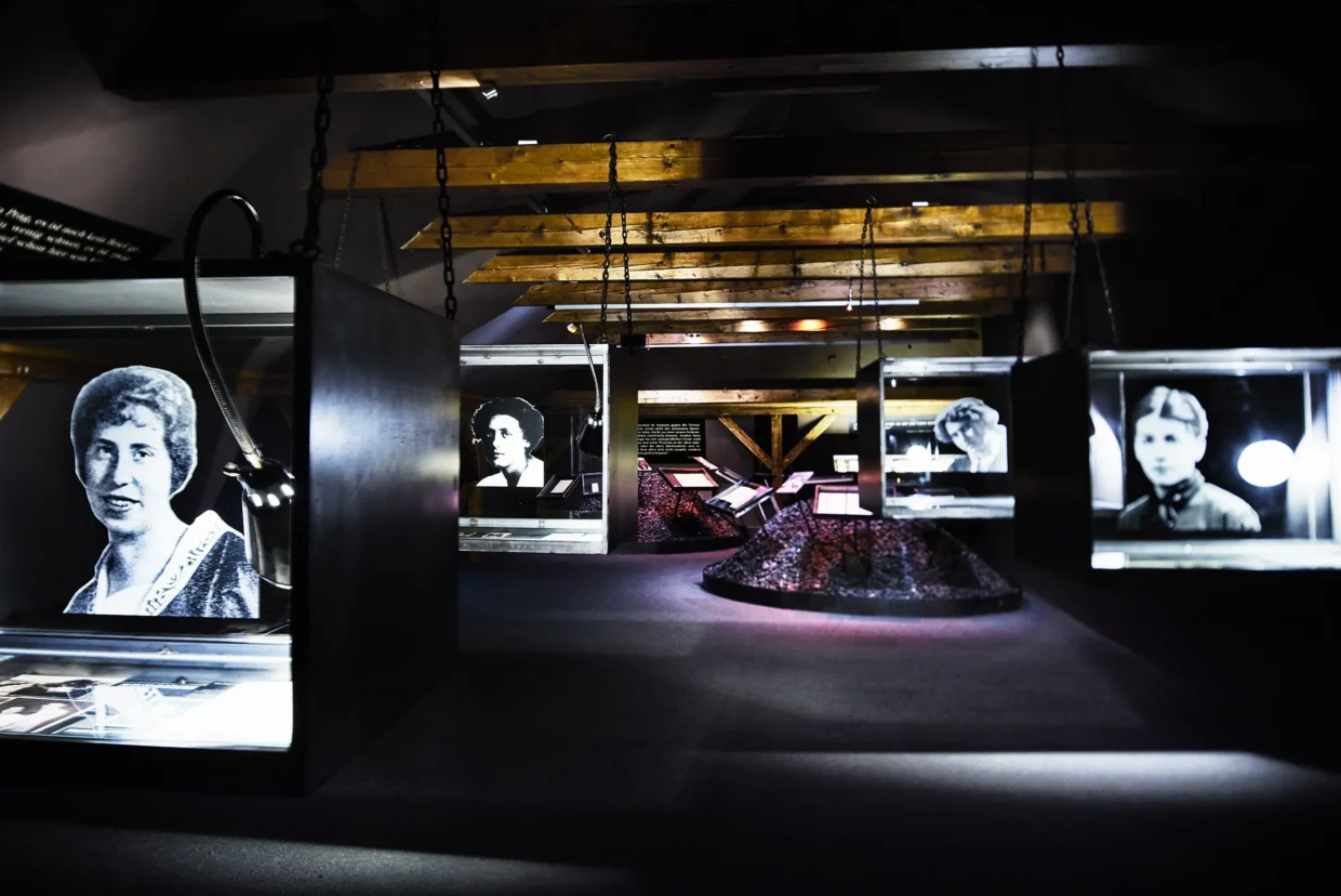 Interior view of the Franz Kafka Museum in Prague, showcasing illuminated display cases with black-and-white portraits and historical documents under wooden ceiling beams.