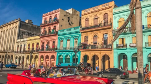 Colorful colonial buildings and a classic red convertible car taxi driving through a lively street in Havana, Cuba, under a clear blue sky.