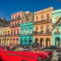 Colorful colonial buildings and a classic red convertible car taxi driving through a lively street in Havana, Cuba, under a clear blue sky.