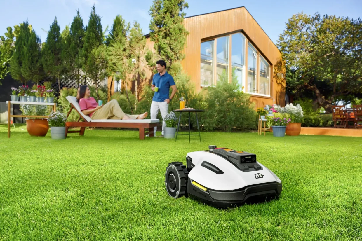 A robotic lawn mower automatically mowing a green backyard, while a woman relaxes on a sun lounger and a man enjoys a drink, with a modern house and lush garden in the background.