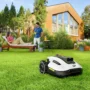 A robotic lawn mower automatically mowing a green backyard, while a woman relaxes on a sun lounger and a man enjoys a drink, with a modern house and lush garden in the background.