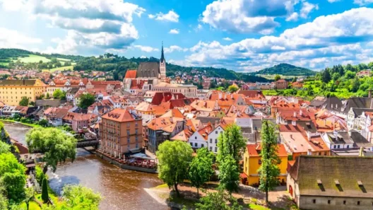 Panoramic view of the historic center of Český Krumlov with red rooftops, the winding Vltava River, St. Vitus Church, and surrounding green landscape under a summer sky.