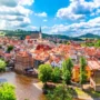Panoramic view of the historic center of Český Krumlov with red rooftops, the winding Vltava River, St. Vitus Church, and surrounding green landscape under a summer sky.