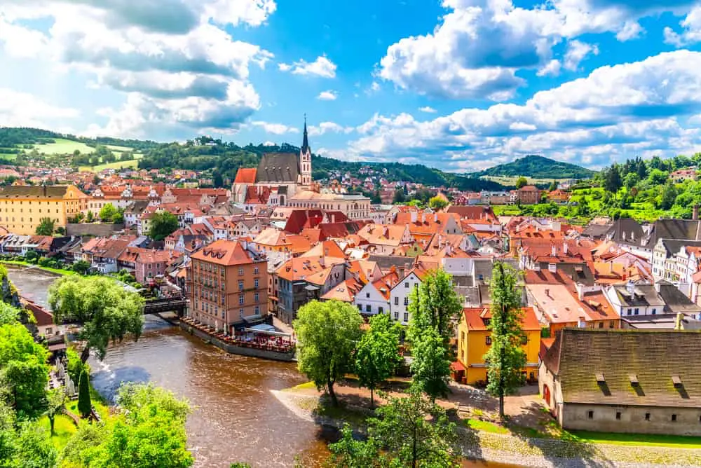 Panoramic view of the historic center of Český Krumlov with red rooftops, the winding Vltava River, St. Vitus Church, and surrounding green landscape under a summer sky.