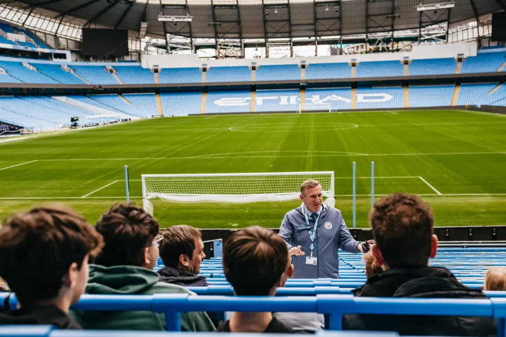 Manchester City stadium tour guide speaking to visitors in the stands at Etihad Stadium, with a view of the empty football pitch and blue seats