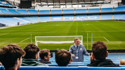 Manchester City stadium tour guide speaking to visitors in the stands at Etihad Stadium, with a view of the empty football pitch and blue seats