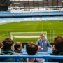 Manchester City stadium tour guide speaking to visitors in the stands at Etihad Stadium, with a view of the empty football pitch and blue seats